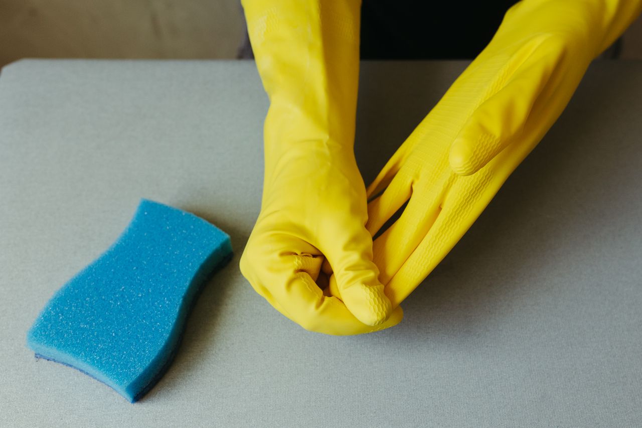 heros-img Close-up of hands wearing yellow gloves next to blue sponge on gray surface.